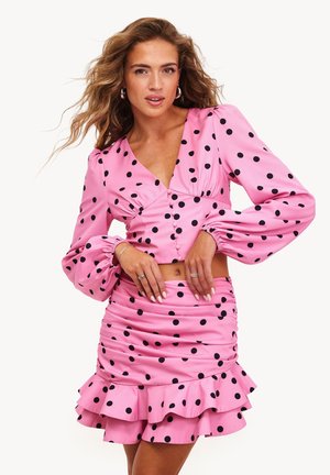 Femme aux cheveux ondulés portant une tenue rose deux pièces à pois noirs, composée d'un crop top à manches longues et d'une mini-jupe à volants.