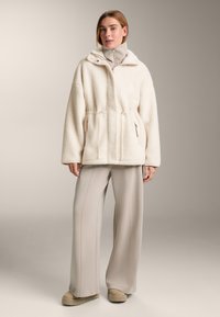 Giacca in pile color crema con cappuccio, vita elasticizzata e due tasche con cerniera. Indossata sopra pantaloni larghi grigio chiaro e scarpe beige con platform.