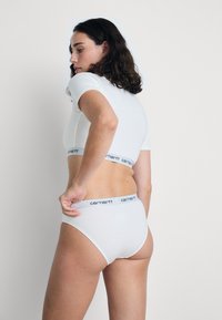 Hvitt crop-top og matchende briefs, begge laget av glatt stoff, har et svart merket liv. Silhuetten er tettsittende og minimalistisk.