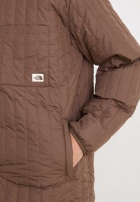 Brązowa pikowana kurtka z logo The North Face na kieszeni na piersi, ręka osoby w kieszeni bocznej, zbliżenie na tors i ramię.