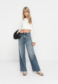 Felpa corta a coste bianca, jeans a gamba larga azzurri chiari e sneakers con motivo. Borsa a tracolla nera. Sfondo luminoso e uniforme.