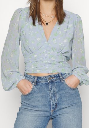 Kvinde med hænder i lommerne iført lyseblå blomstret langærmet bluse med rynket talje og blå højtsiddende jeans, accessoriseret med lagdelte halskæder.