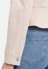 Růžová bunda s dlouhým rukávem a stříbrným drukem, v kombinaci s modrými džínami, které mají zadní kapsu s charakteristickým tvarem a prošíváním.