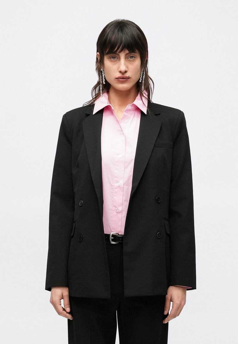 Μαύρο διπλόπετο σακάκι πάνω από ένα ανοιχτό ροζ πουκάμισο με κουμπιά. Το ντύσιμο έχει μακριά μανίκια και διακριτική λάμψη στο ύφασμα.