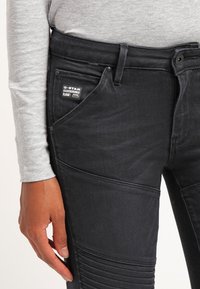 Vaqueros de mezclilla negra con acentos texturizados en las rodillas y un bolsillo lateral, con una etiqueta de G-Star Originals. La tela parece resistente y suave.