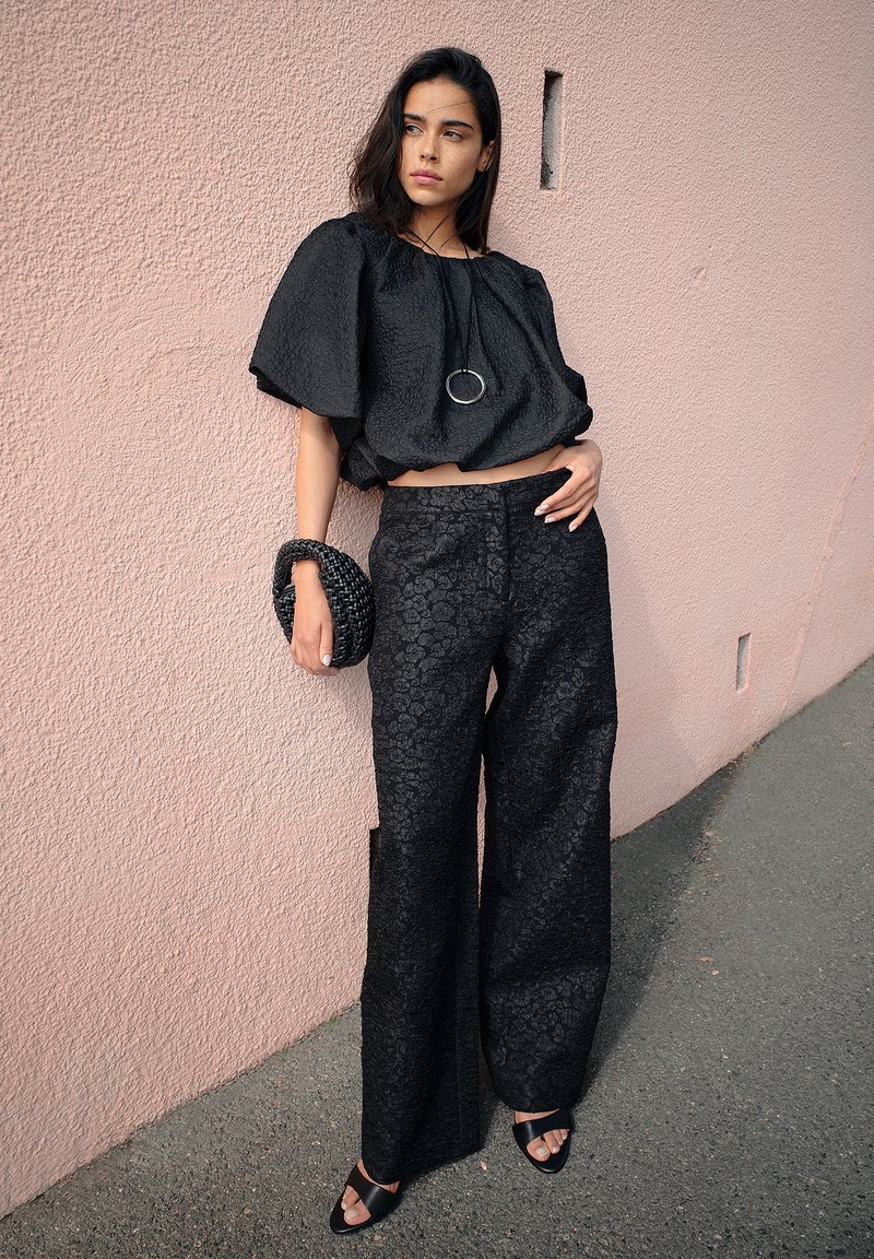 Black textured crop top with loose sleeves, paired with wide-leg black pants featuring a floral pattern. Woven black handbag and black sandals.