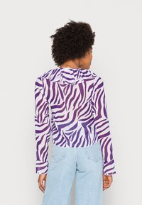 Blusa de manga comprida em tecido transparente com riscas de zebra roxas e brancas, apresentando um colarinho frisado e punhos com botões.