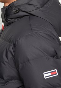 Sort polstret jakke med høj krave, snoretræk i hætten og logo-patch på ærmet. Glat, vandafvisende stof med kviltet tekstur.