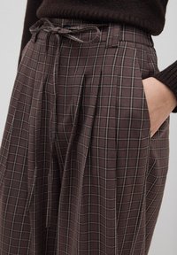Pantalones de cuadros marrones con cintura ajustable, que cuentan con bolsillos laterales y una textura de tela suave. El patrón incluye líneas delgadas en color beige.