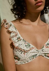 Květinový bikiny top s bílým základem, vzory v zelené a růžové barvě, volánkové ramínka zdobená oranžovými korálky a detail uzlu uprostřed.