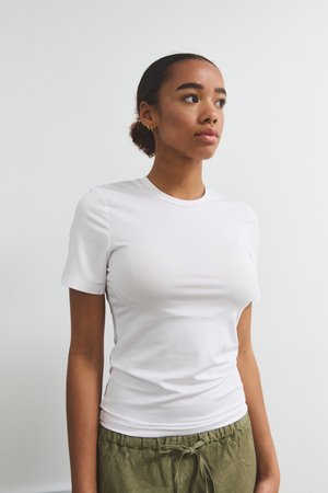 Sieviete ar tumšiem viļņotiem matiem, sasietiem atpakaļ, valkājot īsām piedurknēm pieguļu baltu T-kreklu un olīvzaļas krāsas bikses ar auklu, skatās uz labo pusi.