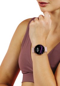 Montre intelligente ronde avec un bracelet rose et des accents métalliques. L'affichage numérique montre l'heure et des éléments graphiques violets. Portée au poignet.