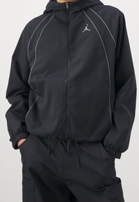 Svart zip-up jacka med huva, med grå detaljer och en Jumpman-logotyp. Tillverkad av ett slätt, lätt material med elastiska ärmslut.