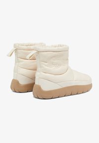 Botas de tobillo acolchadas color crema con una tela texturizada, que cuentan con un detalle de logotipo y una suela de goma marrón con un patrón estriado.