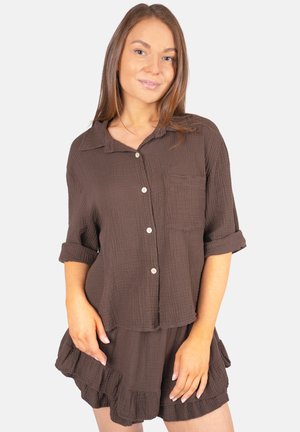 Femme portant une chemise boutonnée marron texturée aux manches retroussées et un short assorti à volants, debout devant un fond clair uni.
