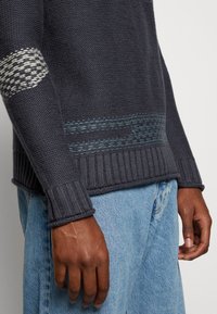 Laivastonsininen neulepaita, jossa on resorireunukset hihansuissa ja helmassa, kevyesti teksturoitu vaaleansininen korostevyö ja tunnistettava kudottu kuvio hihassa.