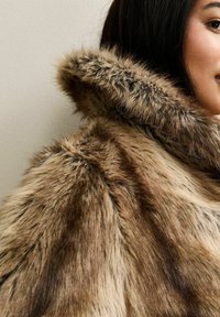 Faux-Fur-Mantel mit einem Farbverlauf von Braun zu Creme, ausgestattet mit einer dicken Kapuze und einer weichen, fusseligen Textur, vor einem schlichten Hintergrund präsentiert.