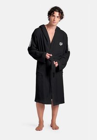 Sort hættebadekåbe lavet af tekstureret stof, med et bundet bælte, sidelommer, et hvidt logo på brystet og mønstrede detaljer på ærmerne.