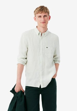 Joven con una camisa clara de rayas y botones, con las mangas remangadas, sosteniendo una chaqueta oscura en una mano, de pie frente a un fondo blanco.