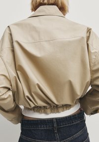 Beige cropped jacket with a soft, textured fabric, featuring a gathered elastic waistband and a classic collar. Back view shows smooth design.