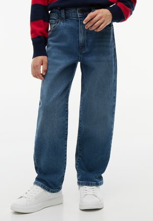 Person trägt lockere blaue Jeans, weiße Sneakers und ein rot-navy gestreiftes Langarmshirt und steht vor einem einfarbigen Hintergrund.