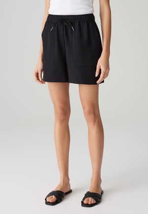 Zwarte, geribbelde shorts met een elastische tailleband en trekkoord. Beschikt over twee voorzakken en een relaxte pasvorm, gecombineerd met zwarte slipper-sandalen.