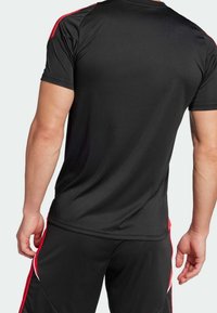 Man i svart träningsoutfit med röd och vit rand, sedd bakifrån, stående mot en enkel ljus bakgrund.