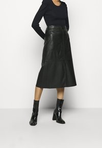 Schwarze Leder-Midi-Rock mit hoher Taille, zwei frontalen Taschen und glatter Textur; kombiniert mit glänzenden schwarzen Absatzstiefeln bis zur Knöchel.