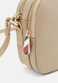 Beige stof crossbody tas med guldfarvet hardware, dobbelt lynlås og en dekorativ charm med røde, hvide og sorte geometriske mønstre.