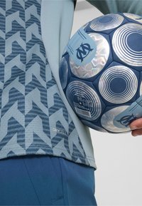 Šviesiai mėlynas sportinis marškinėlis su geometriniu raštu, derinamas su tamsiai mėlynomis šortais. Vienoje rankoje laikomas mėlynas futbolo kamuolys su apvaliais raštais.