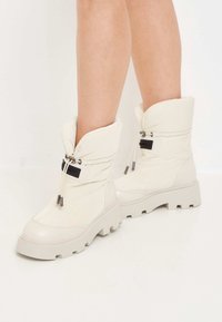 Białe, wypełnione botki za kostkę o pikowanej fakturze, z przednim zamkiem błyskawicznym, czarnymi akcentami i masywnymi szarymi podeszwami.