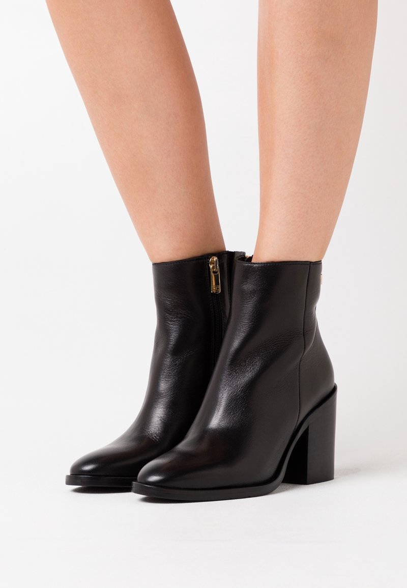 Bottes cheville en cuir noir avec talons blocs et fermetures éclair latérales portées sur des jambes nues sur fond blanc uni