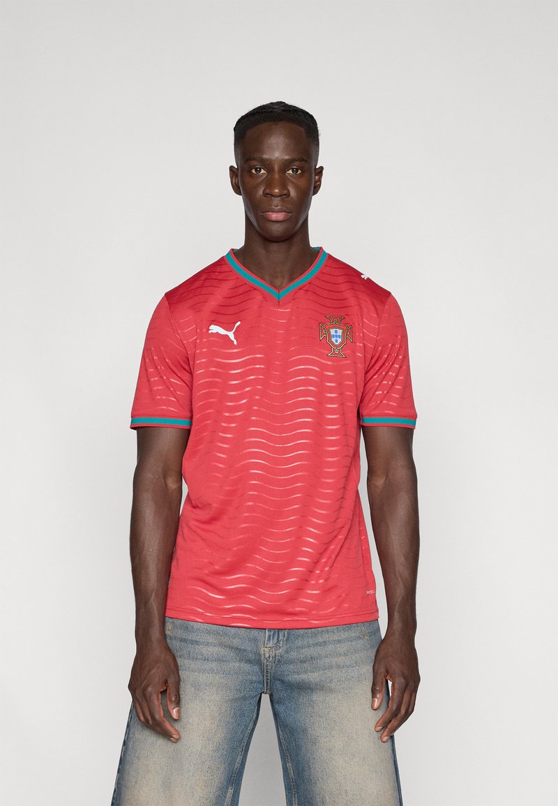 Κόκκινη ποδοσφαιρική φανέλα με κοντά μανίκια και κυματιστό σχέδιο, μπλε λεπτομέρειες στο V-λαιμόκοψη και έμβλημα στο στήθος. Κατασκευασμένη από ελαφρύ ύφασμα.