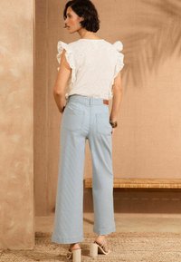 Witte blouse met gerimpelde mouwen, gecombineerd met lichtblauwe wijduitlopende broek met achterzakken en een bruine leren patch. Beige sandalen met blokhak.