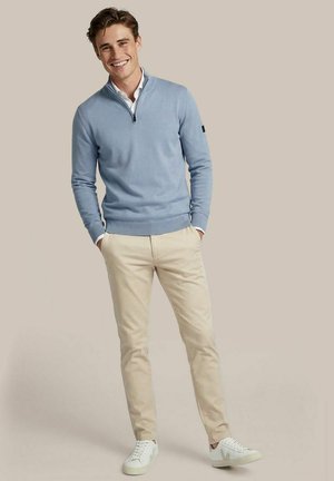 Lachende man staat met handen in de zakken, gekleed in een lichtblauwe trui met kwartrits, wit overhemd, beige broek en witte sneakers, tegen een neutrale achtergrond.