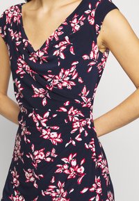 Blommig klänning med marin bakgrund, prydd med rosa och vita blommönster, V-ringning, korta ärmar och en draperad frontdetalj.