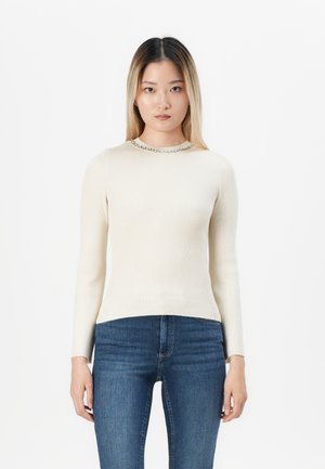Donna con capelli castano chiaro lisci indossa un top a coste crema a maniche lunghe con scollo a gioiello e jeans attillati blu, in piedi su uno sfondo bianco.