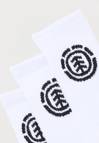 Trois chaussettes blanches côtelées avec un logo circulaire noir présentant un dessin stylisé d'arbre, disposées en chevauchement sur un fond clair.