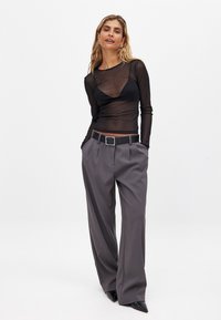 Transparent svart långärmad topp ovanpå en svart bralette, kombinerad med höga grå plisserade byxor och ett svart bälte med fyrkantig spänne.