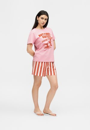 Femme debout portant un t-shirt rose avec un dessin de cafetière et un texte rouge, un short rayé vertical rouge et blanc, et des tongs blanches.