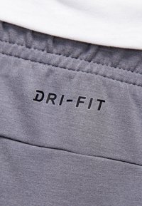 Gros plan d'une ceinture en tissu gris avec le texte noir « DRI-FIT », montrant des coutures élastiques et une partie d'un vêtement blanc au-dessus.