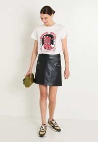 Mini falda de piel sintética negra con cremalleras frontales, combinada con una camiseta gráfica blanca que presenta un diseño de botas coloridas, y un clutch verde oliva.