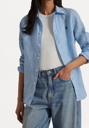 Femme portant une chemise rayée bleu clair et blanc à boutons par-dessus un débardeur blanc côtelé, associée à un jean bleu taille haute, une main dans la poche.