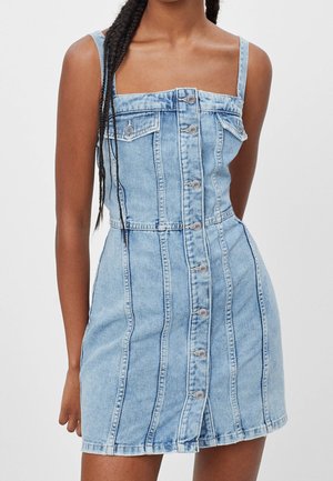 Kobieta ubrana w bezrękawnikową, jasnoniebieską sukienkę dżinsową z zapięciem na guziki z przodu i dwoma kieszeniami na piersiach, stojąca na jednolitym tle.