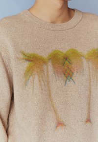 Pull beige en maille avec une texture douce et un motif coloré de palmier. Encolure ronde et bord-côte visibles.