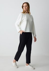 Weißes Oberteil mit langen Ärmeln und kleinen silbernen Akzenten, kombiniert mit schwarzen schmalen Hosen und weißen Turnschuhen, die ein lässiges, stimmiges Outfit hervorheben.