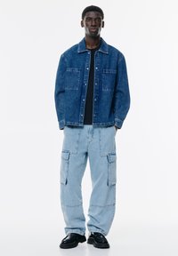 Denim jakna v temno modri barvi z dvema sprednjima žepoma, prekrita s črno majico. Svetlo modri cargo hlače, ki imajo stranske žepke in širok kroj. Črne čevlje.
