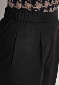 Close-up van zwarte elastische broek met een tailleband, gecombineerd met een beige en zwarte shirt met patroon.