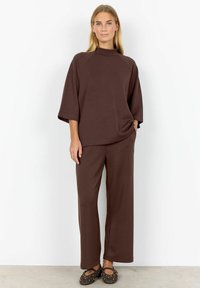 Braunes Loungewear-Set, bestehend aus einem hochgeschlossenen Oberteil mit weiten Ärmeln und lockeren Hosen. Glattes Material mit weicher Textur. Flache Schuhe mit goldenen Nieten.