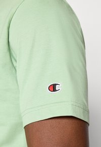 Hellgrünes T-Shirt mit geripptem Textur und kurzen Ärmeln, ausgestattet mit einem kleinen gestickten Logo auf dem linken Ärmel in Rot, Schwarz und Weiß.